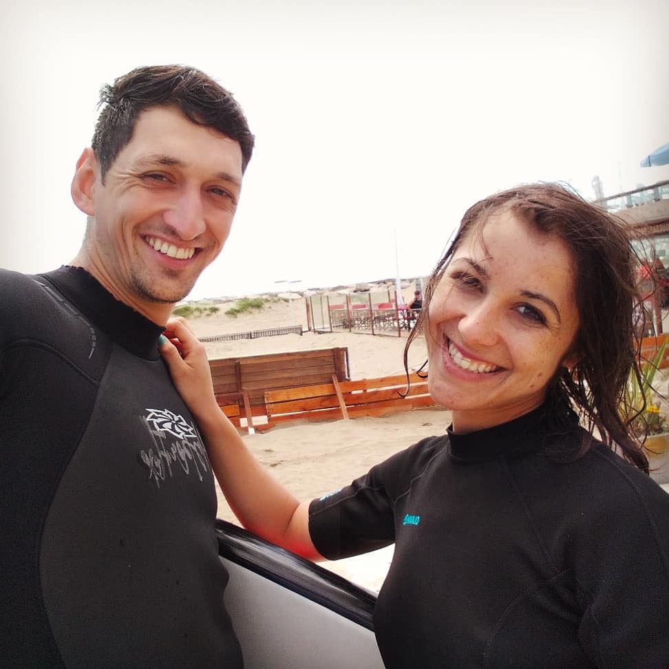 pareja feliz en mar del plata al terminar su sesión de surf, con una longboard y neopreno negro